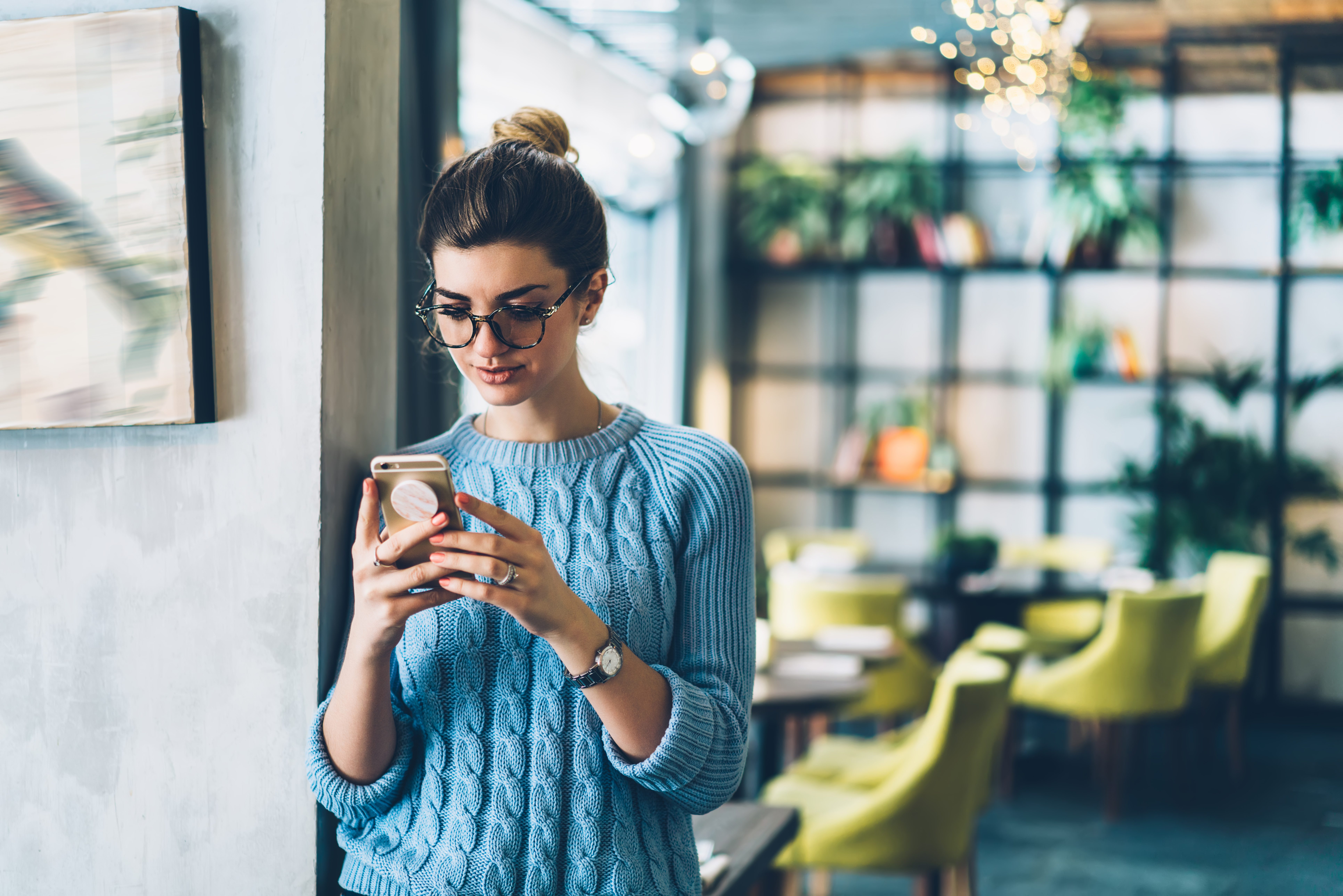 Female blogger chatting with followers in social networks on modern smartphone using 4G internet connection.Hipster girl in stylish eyeglasses watching video on website on cellular standing in cafe